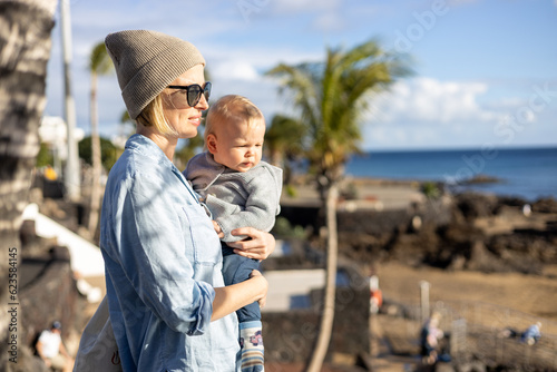 Fashinable mother holding her infant boy child in lap enjoing panoramic view along palm lined beach promenade of Puerto del Carmen, Lanzarote, Canary islands, Spain photo