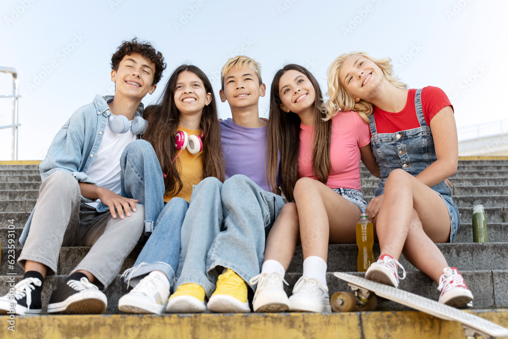 Obraz premium Group of smiling friends, multiracial teenagers wearing colorful t shirts hugging, sitting on stairs outdoors. Happy stylish boys and girls on street. Friendship, positive lifestyle, summer concept 