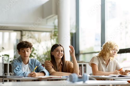 Smart smiling teenage student raising hand answering question sitting in classroom. Group of teenagers studying together  learning language  exam preparation in modern library. Education concept