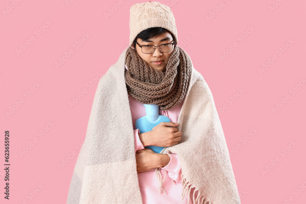 Ill Asian man with hot water bottle on pink background Ill Asian man with hot water bottle on pink background