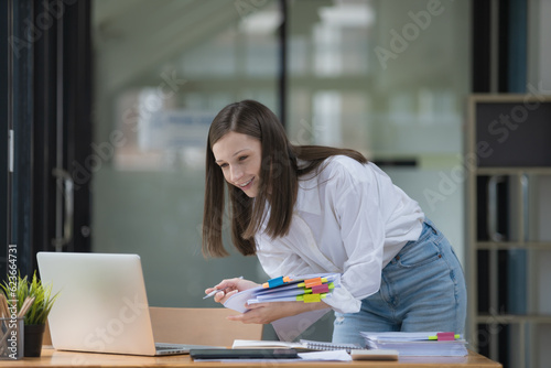 Businesswoman holding a pile of paperwork and using a laptop at her desk in an office. © Songsak C