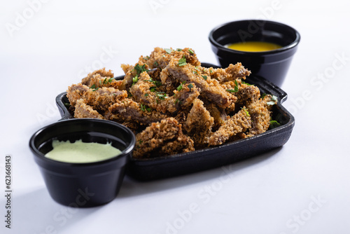 Porção de filé de carne empanada e frito, servida em uma cumbuca preta com molho, em um fundo branco photo