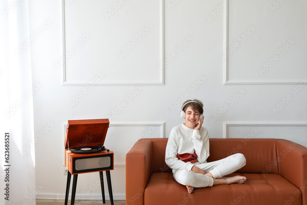 Beautiful happy teenager boy sitting on brown sofa, listening vinyl record on retro wooden turntable, smiling, enjoying music, having fun. Empty white wall background Beautiful happy teenager boy sitting on brown sofa, listening vinyl record on retro wooden turntable, smiling, enjoying music, having fun. Empty white wall background
