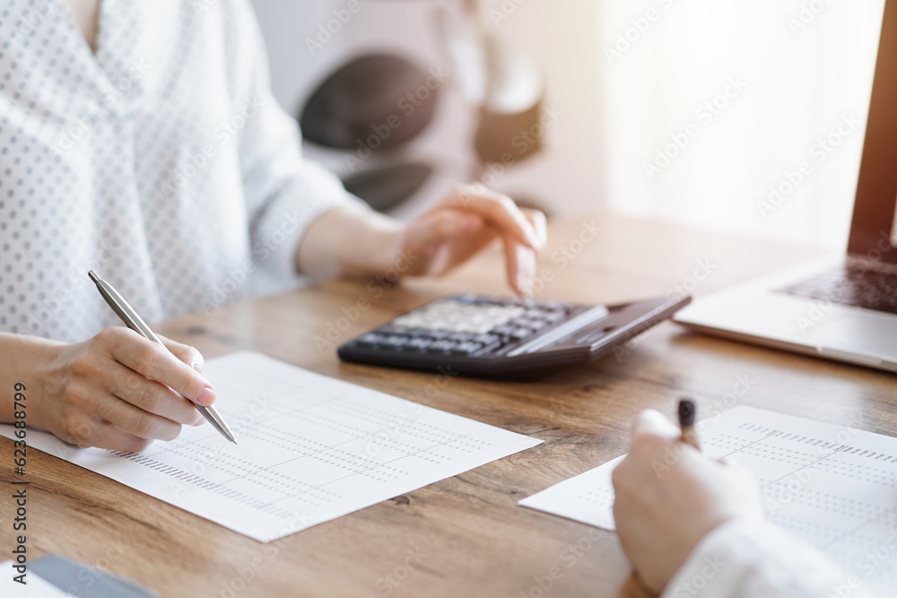Woman accountant using a calculator and laptop computer while counting taxes with colleague at wooden desk in office. Teamwork in business audit and finance Woman accountant using a calculator and laptop computer while counting taxes with colleague at wooden desk in office. Teamwork in business audit and finance