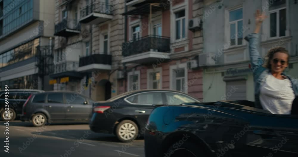 A glossy gray convertible is driving down the street, a blonde girl in the back seat looks out of the convertible and stretches her arms up A glossy gray convertible is driving down the street, a blonde girl in the back seat looks out of the convertible and stretches her arms up