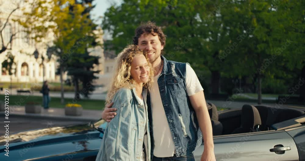 A young beautiful couple of a brunette guy and a blonde girl are standing, posing and looking at the camera against the background of their gray cabriolet on a summer evening A young beautiful couple of a brunette guy and a blonde girl are standing, posing and looking at the camera against the background of their gray cabriolet on a summer evening