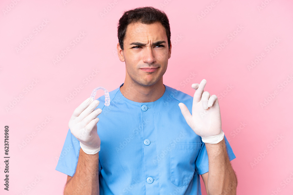 Dentist caucasian man holding invisible braces isolated on pink background with fingers crossing and wishing the best Dentist caucasian man holding invisible braces isolated on pink background with fingers crossing and wishing the best