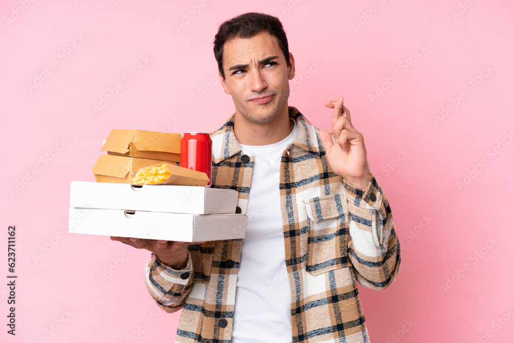 Young caucasian man holding fast food isolated on pink background with fingers crossing and wishing the best Young caucasian man holding fast food isolated on pink background with fingers crossing and wishing the best