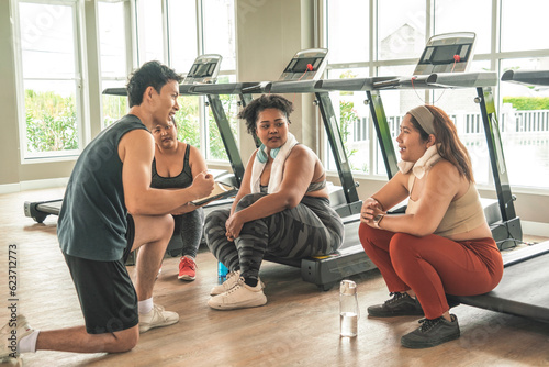 Three plus size women in sports bras sitting on treadmill having fun together and male trainer discussing weight loss and exercising in the gym.