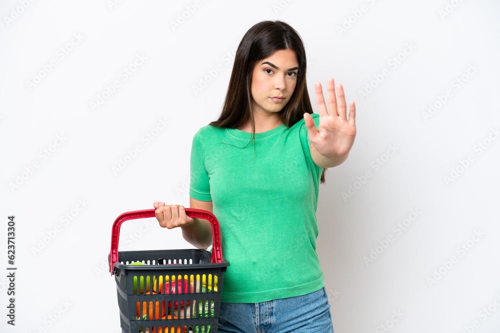 Young Brazilian woman holding a shopping basket full of food isolated on white background making stop gesture Young Brazilian woman holding a shopping basket full of food isolated on white background making stop gesture