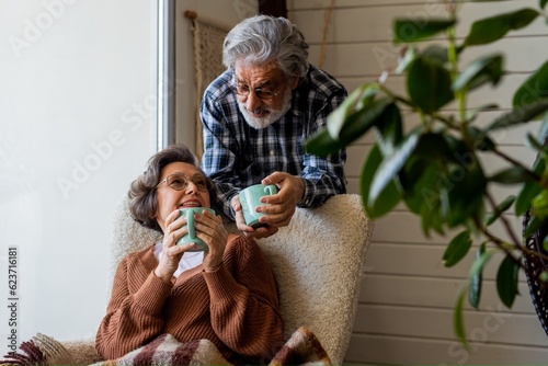 Happy elderly couple relax during holydays. Husband and wife of mature age look gently at each other in a cozy room with cup of coffee in hands