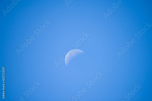 Wieczorne, bezchmurne, niebieskie niebo. W środku kadru widać białą tarczę księżyca. Księżyc wchodzi w fazę ostatniej kwadry. photo