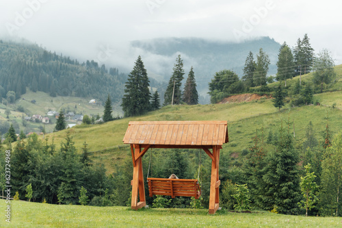A wooden swing stands on a mountain slope with a view of several mountains of Ukrainian Carpathians