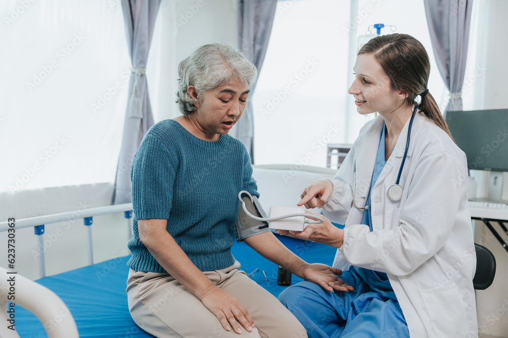 An elderly female patient smiling happily in a light blue dress in bed while a beautiful doctor in a white suit measures blood pressure and checks her pulse in a nursing room. An elderly female patient smiling happily in a light blue dress in bed while a beautiful doctor in a white suit measures blood pressure and checks her pulse in a nursing room.