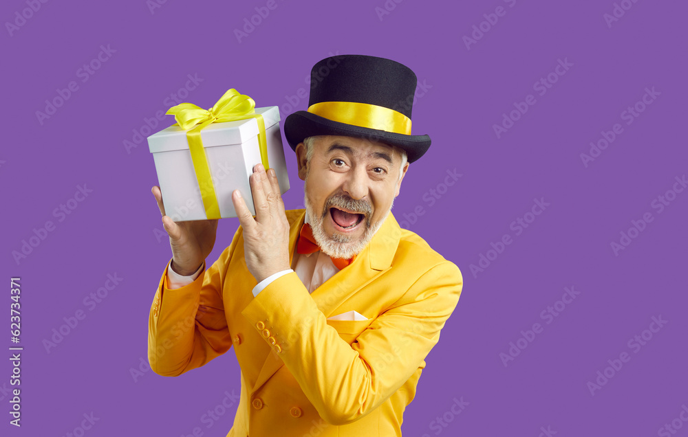 Portrait of overjoyed mature grey-haired man in eccentric yellow suit and hat isolated on purple background hold present. Smiling old bearded male laugh make surprise with promotion gift box. Portrait of overjoyed mature grey-haired man in eccentric yellow suit and hat isolated on purple background hold present. Smiling old bearded male laugh make surprise with promotion gift box.
