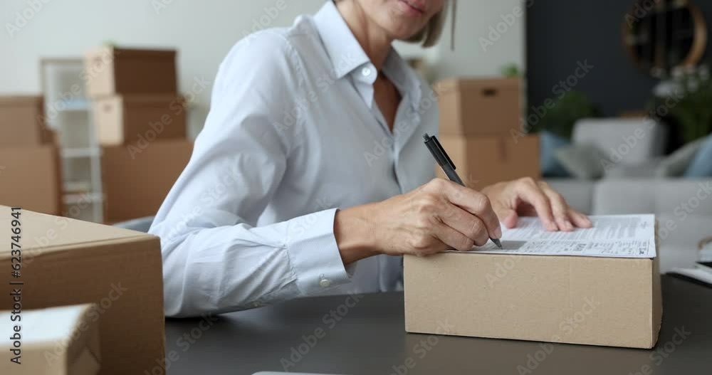 Happy online shop storage manager filling up parcel card, writing in paper document on cardboard box, packaging purchase for customer, preparing package for shipping. Cropped close up shot