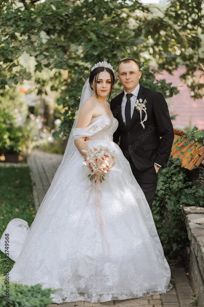 portrait of a young beautiful wedding couple in the garden, the bride in an off-the-shoulder wedding dress, the groom in a black classic suit portrait of a young beautiful wedding couple in the garden, the bride in an off-the-shoulder wedding dress, the groom in a black classic suit