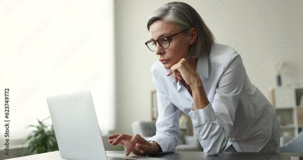 Serious middle aged business woman in glasses working at laptop at home, browsing Internet, watching content, turning look away, thinking on project idea, toughing chin Serious middle aged business woman in glasses working at laptop at home, browsing Internet, watching content, turning look away, thinking on project idea, toughing chin