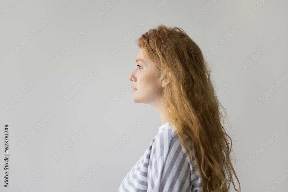 Serious thoughtful young woman with long wavy red hair side view portrait. Pensive pretty girl standing at white wall background, looking away, thinking, dreaming Serious thoughtful young woman with long wavy red hair side view portrait. Pensive pretty girl standing at white wall background, looking away, thinking, dreaming