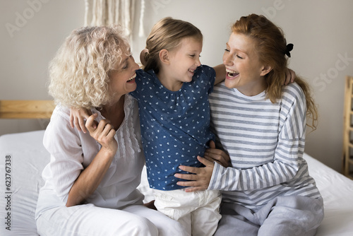 Cheerful little child, mom and grandma having fun on home bed, hugging with love, tenderness, talking, chatting, laughing, enjoying family bonding, funny leisure time