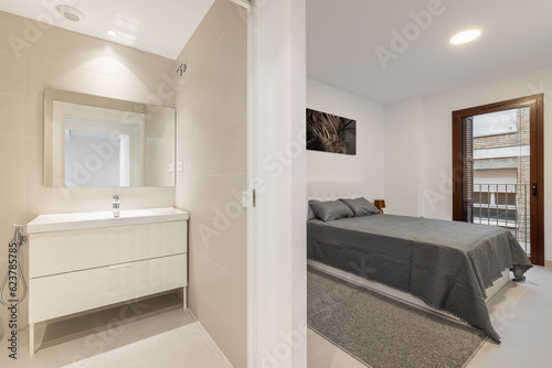 Light, minimalist bathroom with sink, cabinet and mirror in the bathroom with an open door to the bedroom with double bed. Concept of comfortable compact apartment after renovation