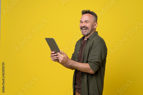 man 50s dressed in green shirt holds digital tablet pc gadget in hands and happy smiled to camera isolated on yellow background