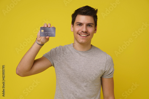 Happy impressed handsome young 20s person holds bank debit card smiling isolated on yellow sunny background