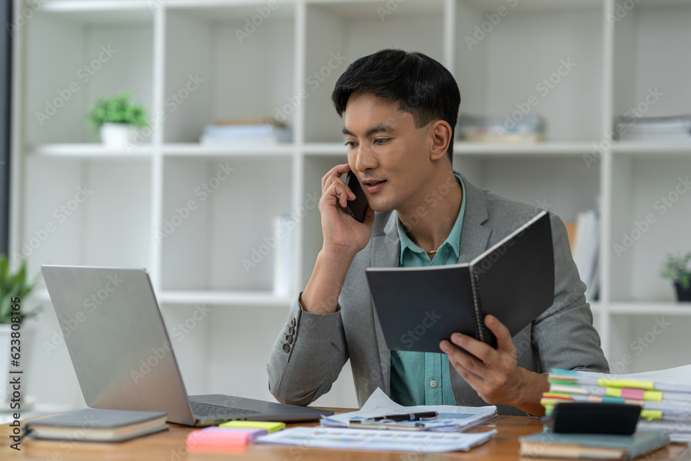 Happy young asian businessman looking at data on laptop, chatting on phone using smartphone for communication. Check through the application Go online while at work in the office. Happy young asian businessman looking at data on laptop, chatting on phone using smartphone for communication. Check through the application Go online while at work in the office.