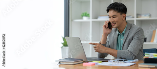 Happy young asian businessman looking at data on laptop, chatting on phone using smartphone for communication. Check through the application Go online while at work in the office.