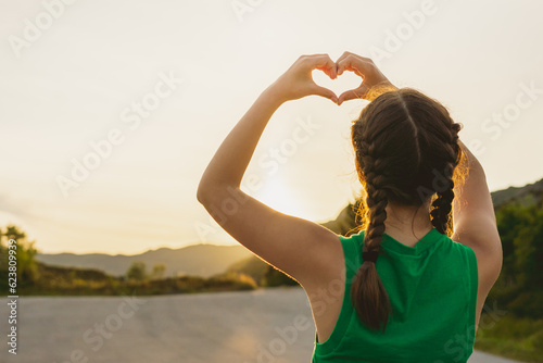 Back view girl in green T-shirt holds her hands in shape of heart against background of sun in nature in mountains