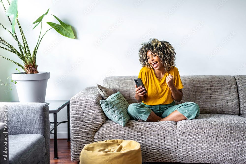 Excited happy young black woman holding smart phone device sitting on sofa at home - Happy satisfied female looking at mobile smartphone screen gesturing yes with clenched fist - Technology concept - obrazy, fototapety, plakaty 