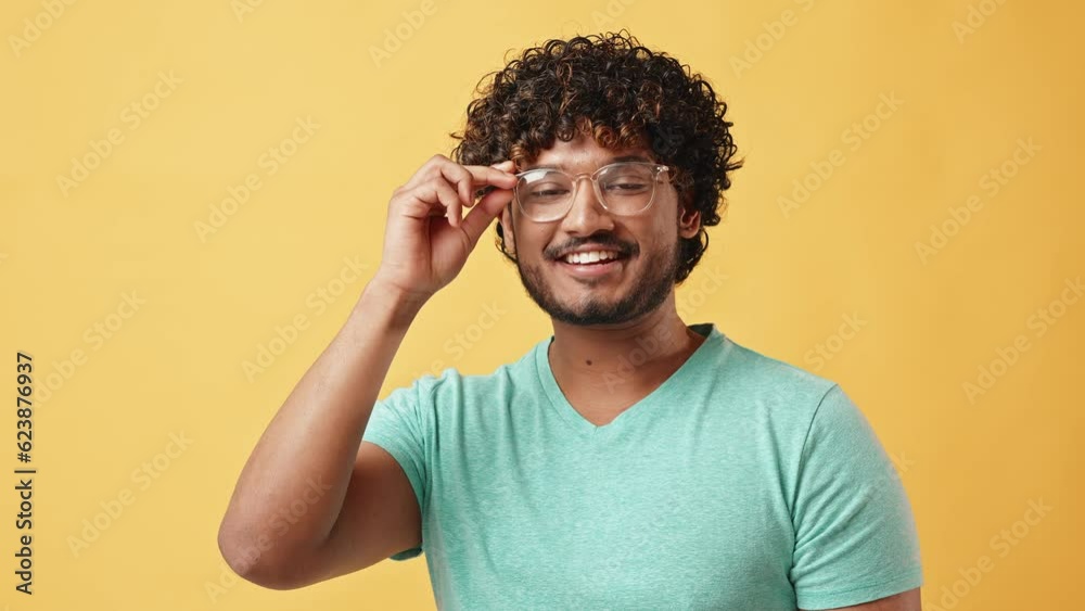 Adult stylish curly modern Indian man in glasses and turquoise t-shirt stands in studio on yellow background and adjusts glasses. Adult stylish curly modern Indian man in glasses and turquoise t-shirt stands in studio on yellow background and adjusts glasses.