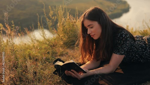 Young christian girl reading the bible, praying, putting her hand on the bible, lying in the grass on the rock in nature with an incredible view, sunset, peace and tranquility, faith