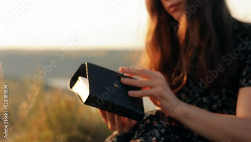 Close-up portrait of an attractive young woman reading a Bible attentively outdoors at sunset. The girl opens the book in a good mood, smiles happily and joyfully
