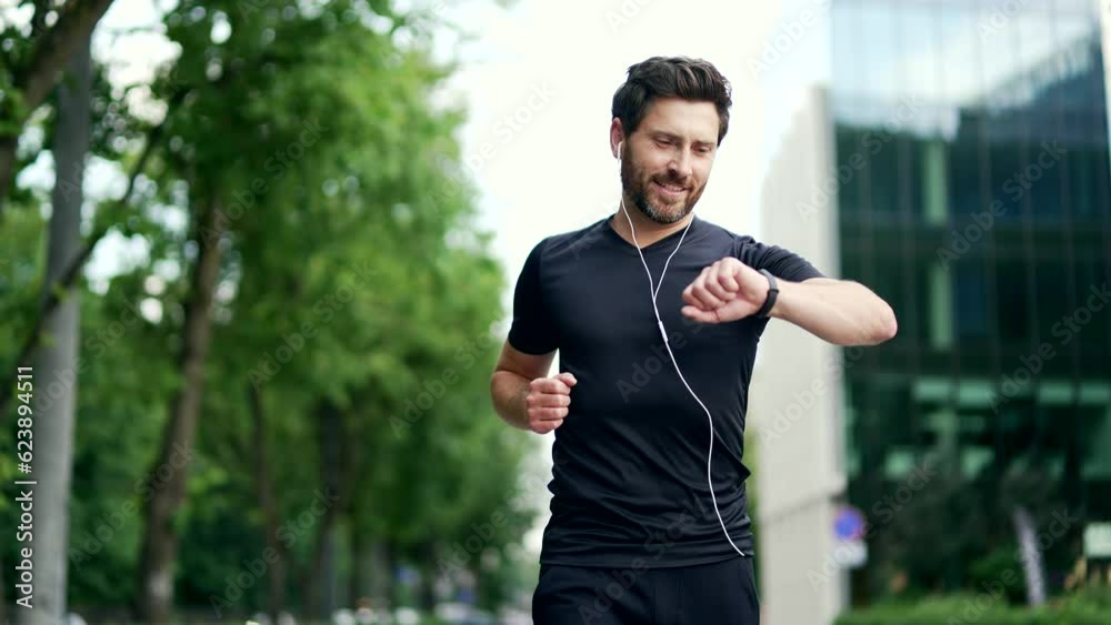Handsome bearded male runner running on a modern urban background of buildings a city street. Sporty man jogger in headphones jogging outdoors look at result a fitness tracker bracelet or smartwatch