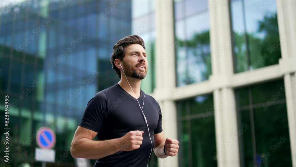 Handsome bearded male runner running on a modern urban background of buildings a city street. Sporty man jogger in headphones jogging outdoors look at result a fitness tracker bracelet or smartwatch