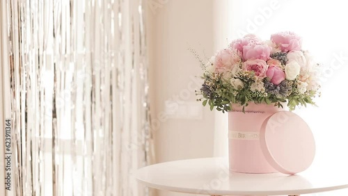 An elegant bouquet of fresh roses, peonies, and daisies in a pink box, beautifully arranged and placed by a light-filled window.