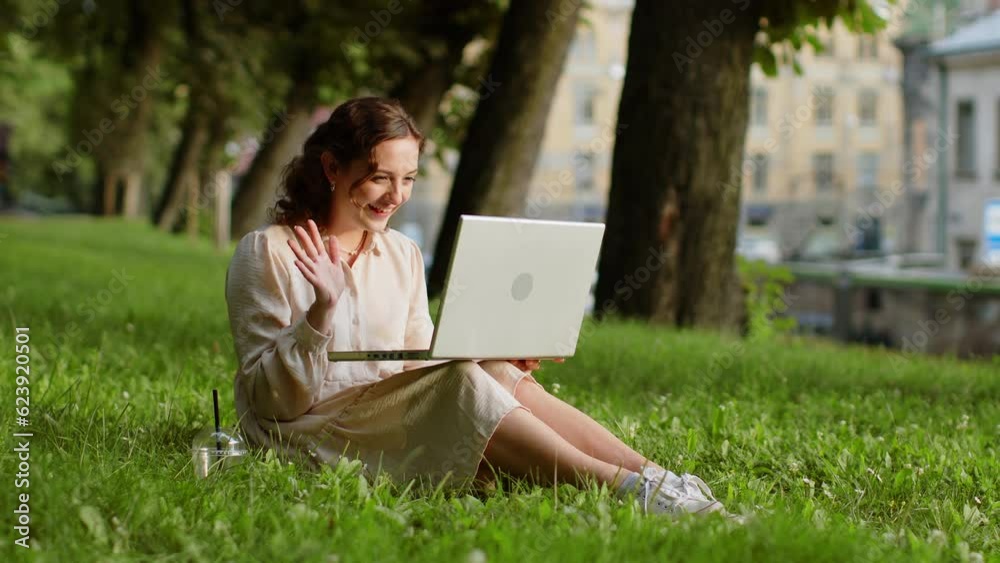 Portrait of pretty young woman making video laptop webcam conference call with friends or family enjoying pleasant conversation, waving hello, laughing. Girl sitting on grass in urban sunset city park Portrait of pretty young woman making video laptop webcam conference call with friends or family enjoying pleasant conversation, waving hello, laughing. Girl sitting on grass in urban sunset city park