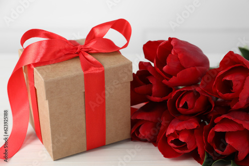 Beautiful gift box with bow and red tulip flowers on white table  closeup