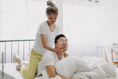 Funny asian couple wife giving husband hurt massage in white bedroom.