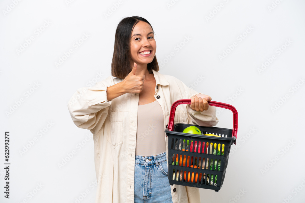 Young woman holding a shopping basket full of food isolated on white background giving a thumbs up gesture Young woman holding a shopping basket full of food isolated on white background giving a thumbs up gesture