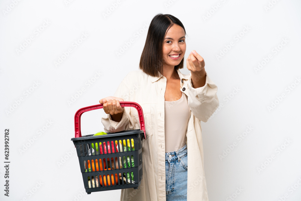 Young woman holding a shopping basket full of food isolated on white background making money gesture Young woman holding a shopping basket full of food isolated on white background making money gesture