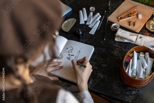 Female artist drawing with ink on white paper, sitting at table with paints, brushes and various art supplies, creating modern artwork in cozy art studio. Talent and creativity