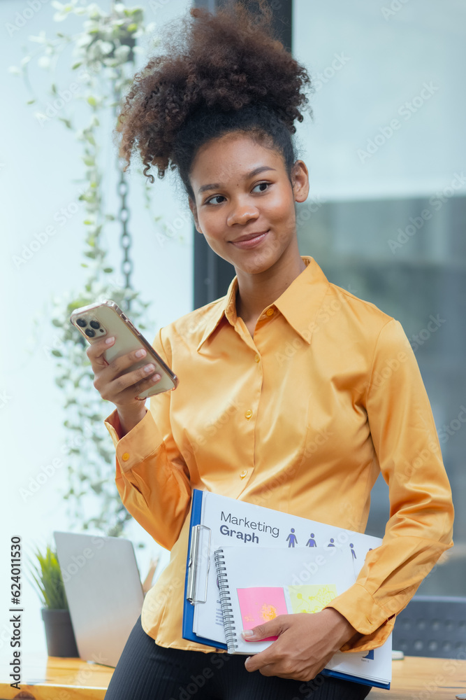 Young African American businesswoman using smartphone with clipboard standing in office. African American businesswoman. Young African American businesswoman using smartphone with clipboard standing in office. African American businesswoman.