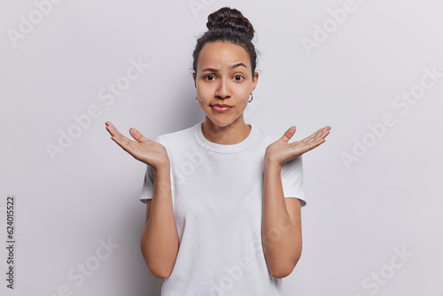Waist up shot of confused Latin woman raises her hands with palms open conveying sense of hesitation doubt unable to make decision makes difficult choice wears casual t shirt isolated on white wall