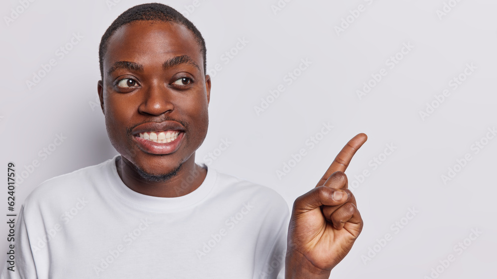 Horizontal shot of dark skinned man smiles toothily points index finger on copy space for your advertisement dressed in casual tshirt isolated over white background. People and promotion concept Horizontal shot of dark skinned man smiles toothily points index finger on copy space for your advertisement dressed in casual tshirt isolated over white background. People and promotion concept