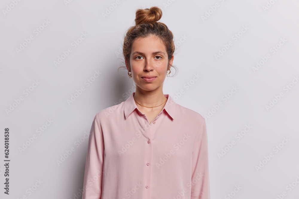 Professional studio portrait of young happy brunette beautiful pierced European lady isolated in centre on white background wearing pink blouse keeping hands down with bun. People emotions concept Professional studio portrait of young happy brunette beautiful pierced European lady isolated in centre on white background wearing pink blouse keeping hands down with bun. People emotions concept
