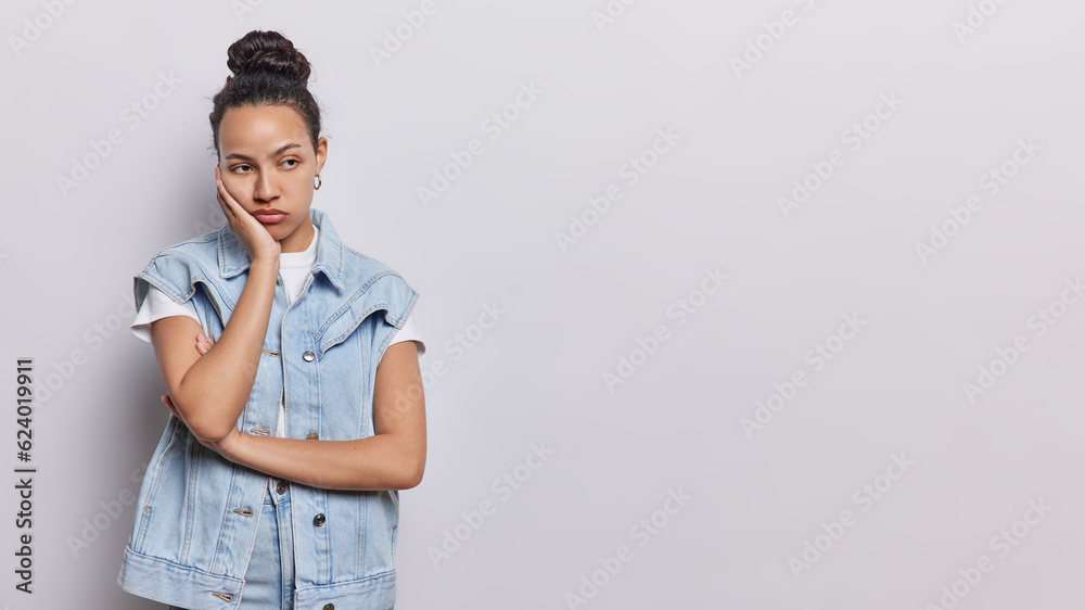 Horizontal shot of displeased Latin woman has sad and dejected expression looks away her eyes filled with frustration and worry keeps hand on cheek dressed in denim vest isolated over white background Horizontal shot of displeased Latin woman has sad and dejected expression looks away her eyes filled with frustration and worry keeps hand on cheek dressed in denim vest isolated over white background