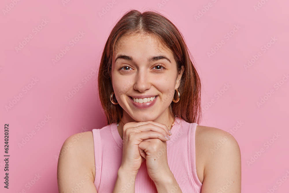 Beautiful smiling broadly young fit European lady with long straight loose brown hair wearing casual tshirt golden earrings and handmade necklace isoalted on pink background holding hands under chin Beautiful smiling broadly young fit European lady with long straight loose brown hair wearing casual tshirt golden earrings and handmade necklace isoalted on pink background holding hands under chin