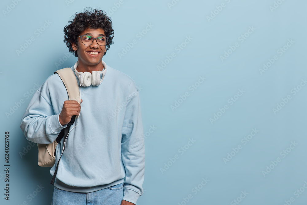 Horizontal studio photo of young delighted smiling Hindu wearing casual blue hoodie and jeans with white bag isolated on left on blue background looking at blank space for your advertisement Horizontal studio photo of young delighted smiling Hindu wearing casual blue hoodie and jeans with white bag isolated on left on blue background looking at blank space for your advertisement